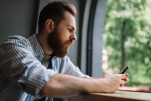 Giovane Hipster Barbuto Con Smartphone Ricerca Informazioni Prenotazione Hotel Seduto — Foto Stock