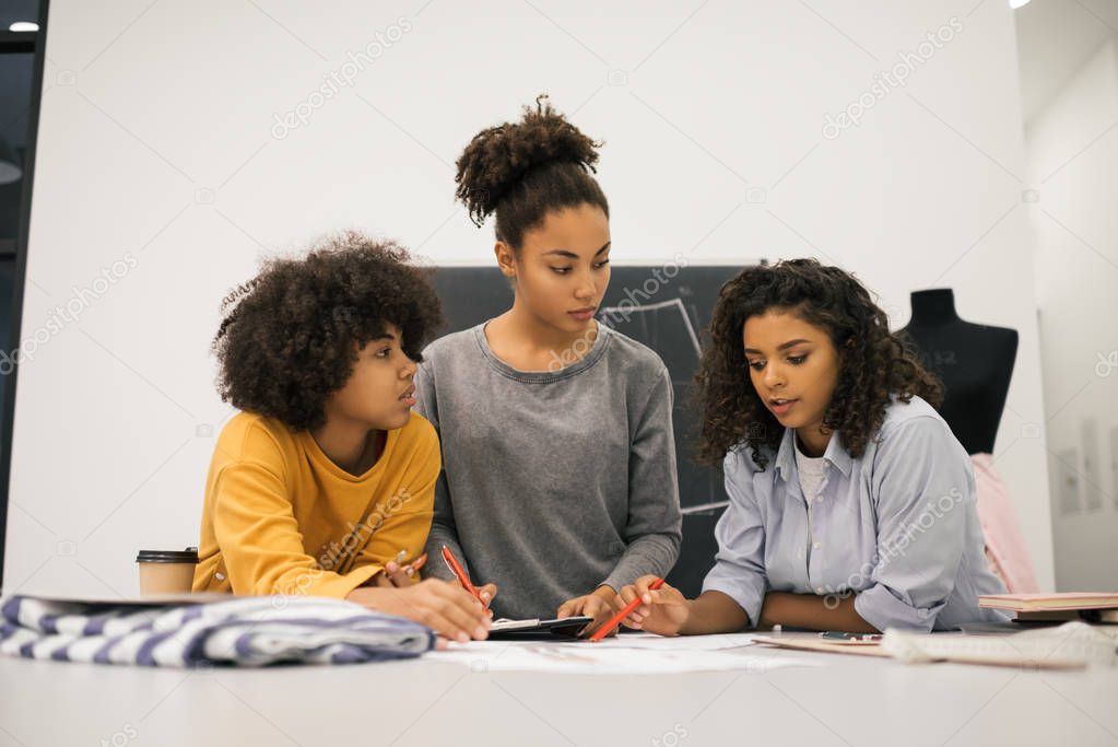 Creative team of stylish African American women planning new startup project in workplace. Brainstorming. University students learning fashion design at atelier. Young hipsters working together.