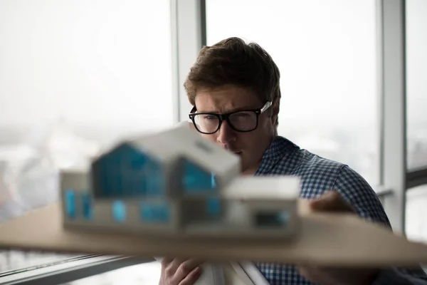 Young architect holding a home model in loft modern office. Architectural project concept. University student wearing stylish glasses planning engineering design at workplace.