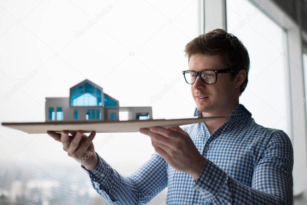 Smiling architect engineer standing near windows in modern loft office. Well dressed student wearing stylish glasses planning new project at university library, holding house model. Education concept.