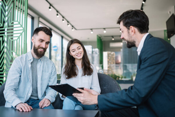 Счастливая молодая пара с улыбчивыми эмоциональными лицами выбирает комфортное жилье в агентстве недвижимости. Покупка дома и собственности вместе. Финансовый консультант в современном офисе
. 