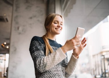 Gülümseyen, pozitif sarışın kadın akıllı telefon tutarak ve modern kafede seyahat online rezervasyon. Hipster kadın güzel yüzünü uygulaması indirme ile. Mobil bankacılık. Nakit geri.