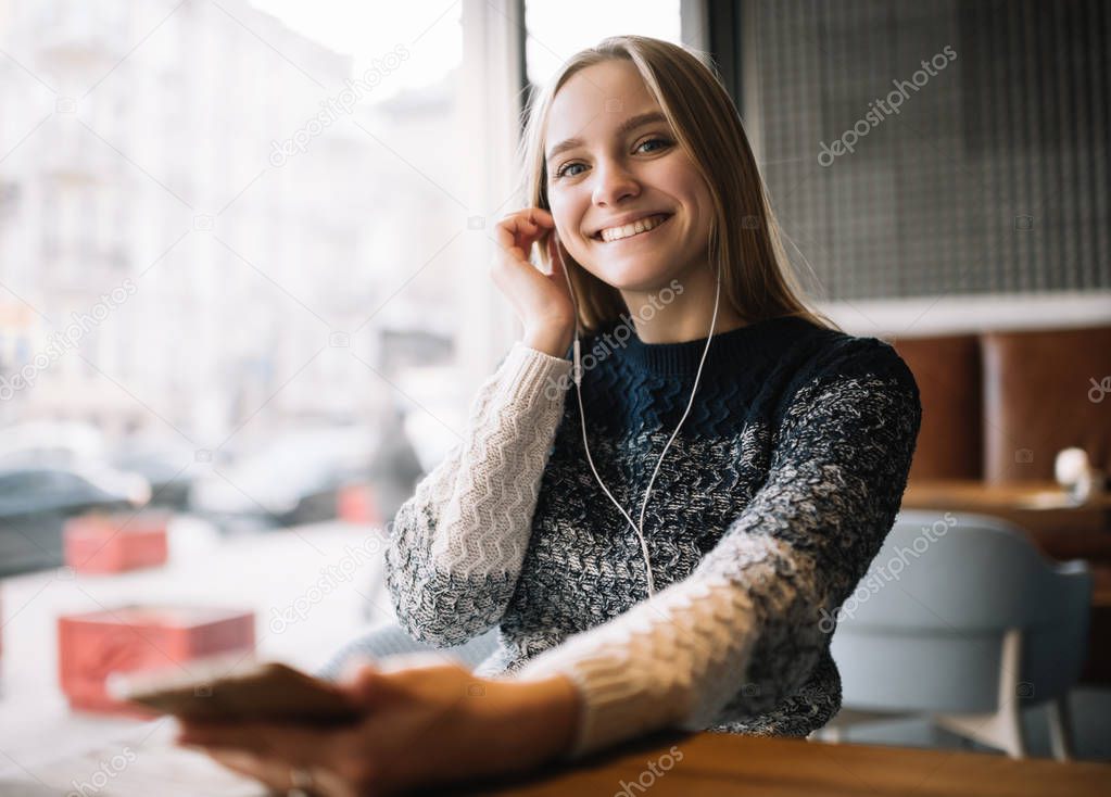 Beautiful smiling woman listen to music, using headphones and smartphone in loft cafe. Young positive female holding mobile phone, enjoying sound of music,  she is in good mood