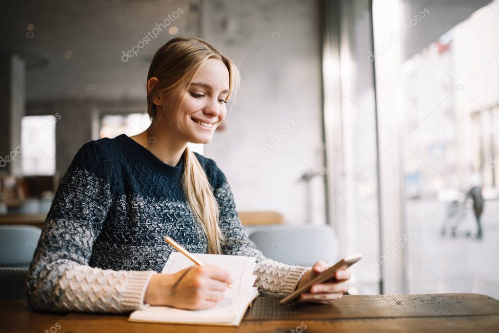 Young attractive university student using laptop computer, studying at modern library. Cheerful caucasian woman writes notes, planning working process, sitting in loft cafe. Exam preparation. 