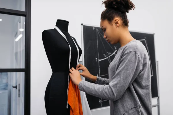 Beautiful dark skinned dressmaker making custom clothing for clients, holding measuring tape, standing near mannequin in tailor shop. African American university student learning fashion design.
