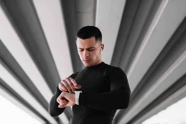 Portrait of muscular athlete listening music wearing wireless headphones and using smartwatch standing on the street . Professional sportsman or runner  in fitted suit ready to workout. Training.