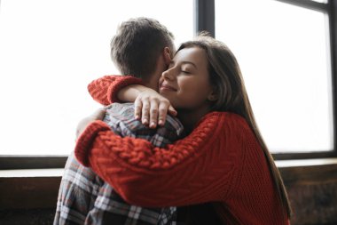 Loft konforlu daireler içinde genç güzel aile portresi. Güzel yüzünü sıcacık evde kocası sarılma kadınla. Çift ayakta birlikte pencere yakınındaki gülümseyen güzel.