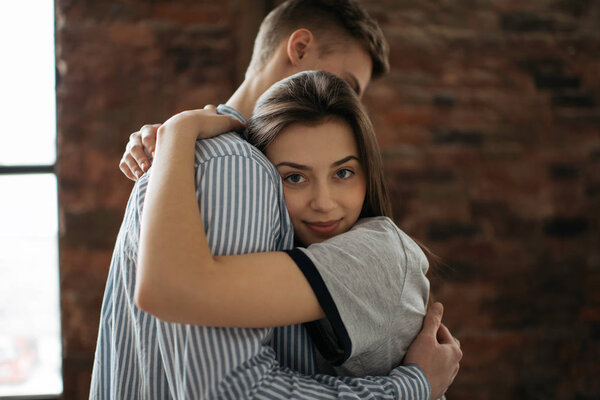 Прекрасная пара обнимается дома, оставайтесь в безопасности. Портрет молодой семьи, стоящей у окна 
