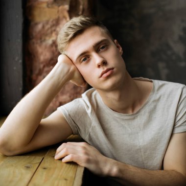 Resimler için poz şık t gömlek giymiş adam çekici mavi gözlü. Çatı katı modern iç penceresinde yakın oturan ciddi şık erkek modeli. 