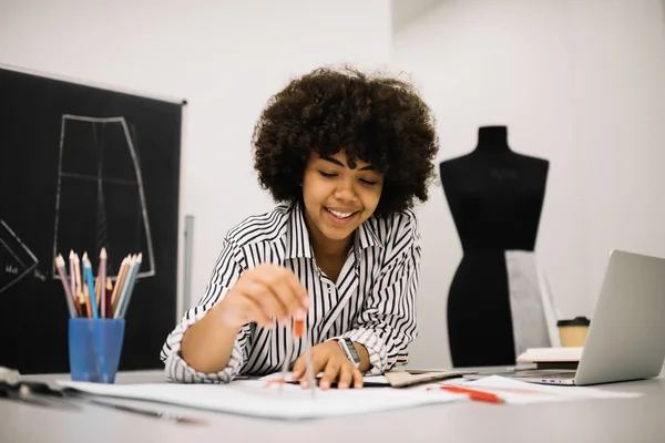 Smiling African American fashion illustrator sketching and drawing at office using drawing compass. Portrait of attractive curly woman working in modern atelier. Freelance.