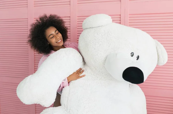 Smiling African American woman with beautiful face and curly hairstyle in pink denim jeans jacket and sexy bodice  hugging large plush  bear toy. Birthday present for positive dark skinned female.