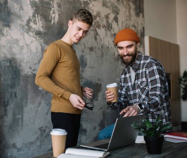 Kahve içme ve dizüstü bilgisayar kullanan loft modern ofis gülümseyen hipsters arkadaşlar. Eğitim, araştırma, uygulama indirme üniversite öğrencileri. Online eğitim kavramı. 