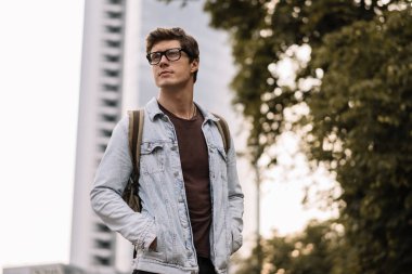 Genç yakışıklı öğrenci üniversiteye gidiş sırt çantası ile. Ciddi yüz kot ceket ve sokakta taksi bekleyen şık hipster gözlük takıyor kendine güvenen adam portresi. 