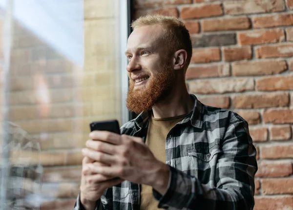 Happy, positive bearded guy using smartphone for booking travel online in modern coffee shop. Hipster man with emotional smiling face downloading application, dreaming, looking at window. Mobile banking. Cash back.