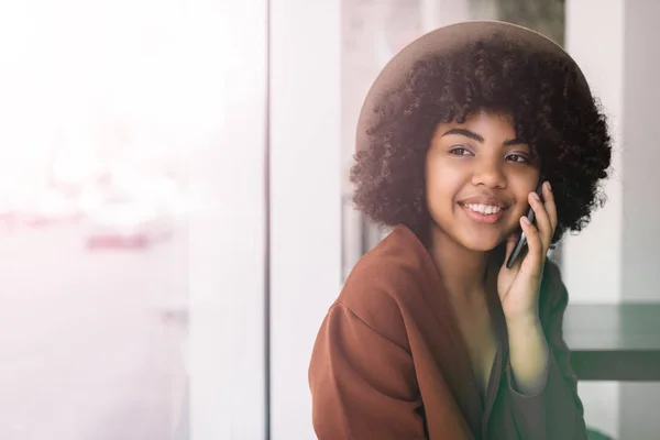 Authentic portrait of smiling African American woman with happy emotional face and curly hairstyle using mobile phone for communication. Hipster girl calling taxi sitting in modern cafe.