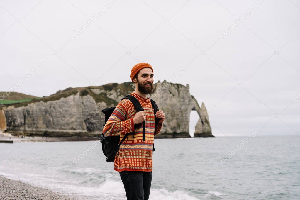 Authentic portrait of happy bearded traveler with smiling emotional face and colorful clothing walking along the beach near sea and cliffs, enjoying the beauty of nature. Bearded hipster man tourist with backpack hiking alone. 