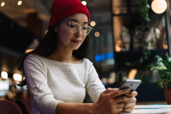 Beautiful asian woman wearing stylish hipster hat and optical lens glasses for best vision watching movie using mobile phone and internet sitting in cafe. Successful blogger streaming video online, chatting, blog posts