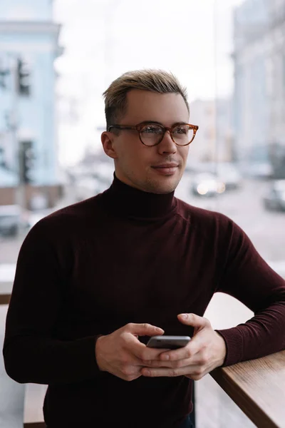 Handsome man using mobile phone sitting in cafe, waiting for taxi. Successful blogger wearing hipster eyeglasses and stylish turtleneck holding smartphone, blog post, texting message, brainstorming, thinking, looking at window.