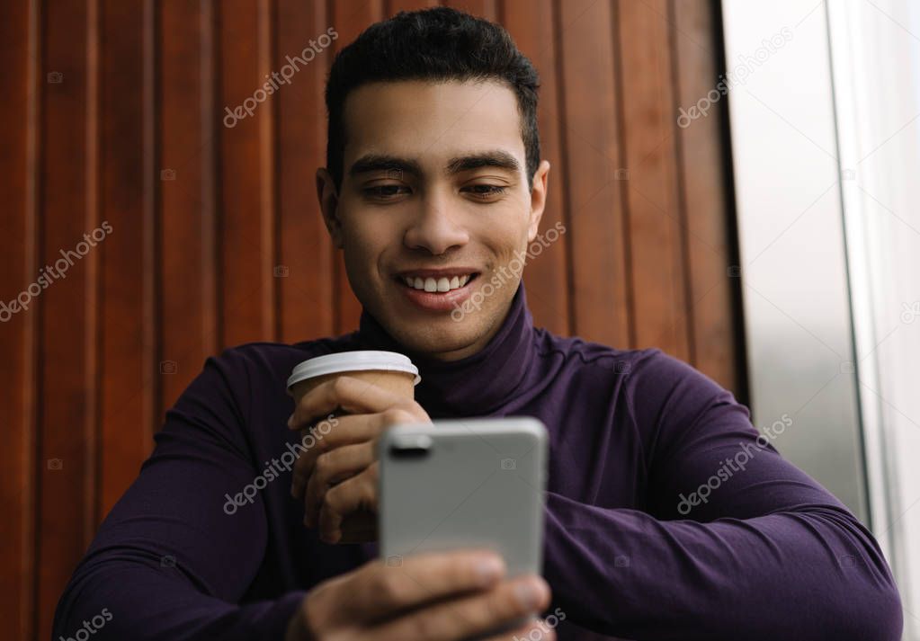Happy African American man using cellphone and mobile application for online shopping with discount sales, cash back, low prices. Smiling blogger blog post, streaming video, chatting, drinking coffee in cafe 