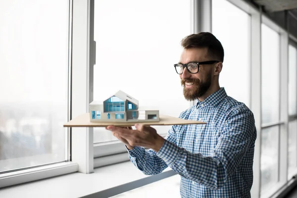 Successful smiling architect holding model house in loft modern office. Portrait of young handsome engineer working project, planning strategy, looking for creative solution at workplace. Smart home concept