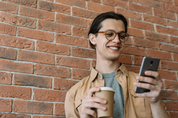 Young smiling man using mobile phone for online shopping, he drinking coffee, standing on the street near brown wall. Portrait of happy emotional hipster in stylish eyeglasses chatting, streaming video online, communication
