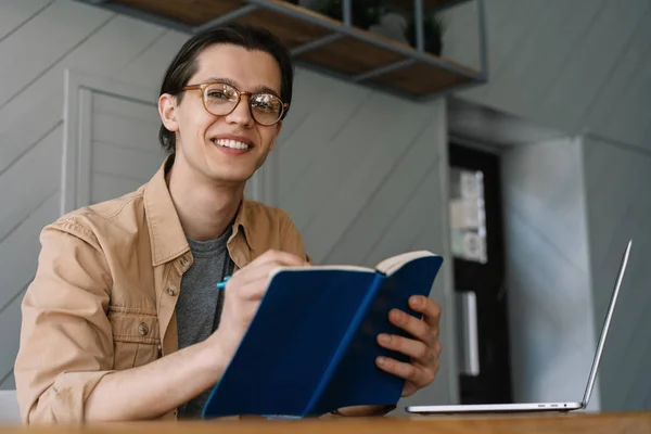 Young handsome university student using laptop computer, studying at library, learning language, exam preparation. Education concept. Smiling hipster man writes notes in notebook working freelance project, planning strategy, brainstorming in cafe