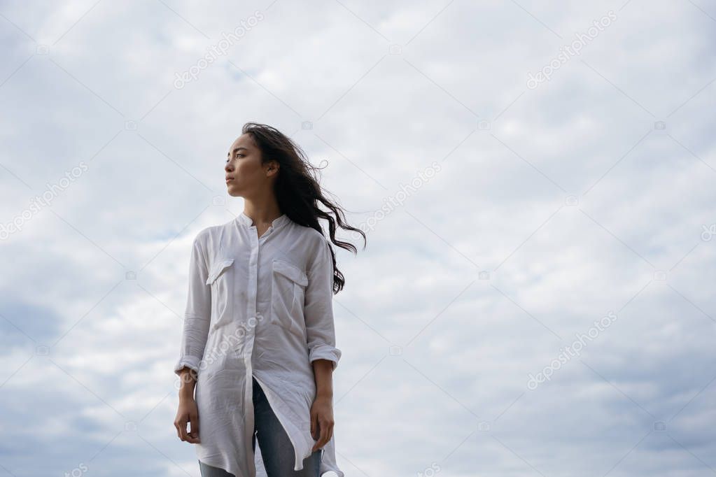 Authentic portrait of christian woman breathing fresh air, thinking, makes important life decisions. Asian girl with emotional face standing on background of clouds. Faith in God. Religion concept 