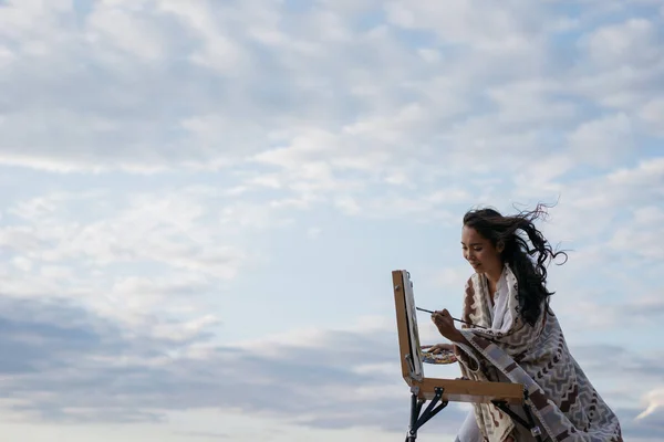 Happy asian artist painting picture on canvas outdoor with clouds on background. Portrait of cheerful Korean woman painter holding paintbrush and colorful palette, admire beautiful landscapes and amazing sunset