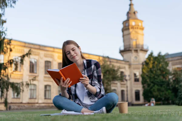 Successful university student studying, reading a book, learning language, exam preparation, sitting on grass. Portrait of young happy woman with emotional face working in park. Education concept