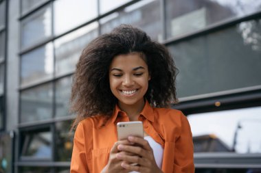 Online alışveriş için mobil uygulama kullanarak neşeli Afrikalı Amerikalı kadın Portresi. Sosyal ağlara Başarılı blogger blog yazısı. Gülümseyen kız akıllı telefon tutan, sohbet, bilet rezervasyonu