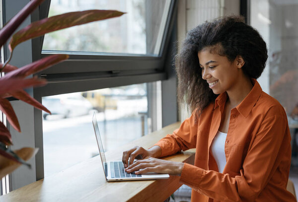 Happy African American woman copywriter working freelance project in modern cafe. Young businesswoman using laptop, searching information on website, typing text, planning strategy, Successful business. Online training courses concept
