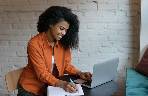 Успешное обучение студентов университета, изучение языка, использование ноутбука и Интернета. Молодая счастливая афроамериканка смотрит онлайн бизнес-курсы на сайте, делает заметки
 
