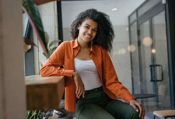 Portrait of female small business owner sitting on chair in modern cafe. Attractive African American woman with happy emotional face, afro hairstyle posing for pictures, laughing