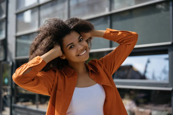 Cute African American fashion model posing for pictures outdoors. Portrait of cheerful curly hair woman with beautiful smile and white teeth walking on street. Hair volume, natural beauty concept