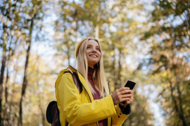 Güz ormanlarında seyrüsefer için mobil uygulama kullanan güzel bir kadın. En iyi yolu bulmaya çalışan mutlu bir turist. Aktif yaşam tarzı, seyahat kavramı