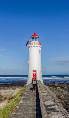 Siyah giyinmiş genç bir kadın beyaz bir deniz fenerine doğru yürüyor, Great Ocean Road, Avustralya