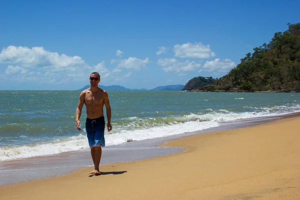 Hombre musculoso caucásico caminando en la playa y sonríe mientras las olas del océano se estrellan detrás de él, la tribulación del cabo, Queensland, Australia — Foto de Stock