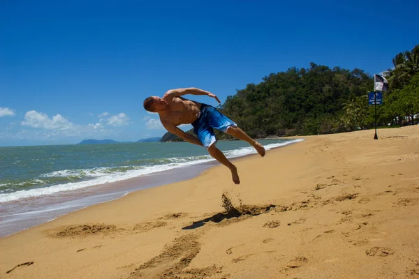 Hombre musculoso caucásico haciendo una voltereta en la playa mientras las olas del océano se estrellan detrás de él, la tribulación del cabo, Queensland, Australia — Foto de Stock