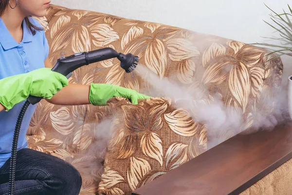 掃除だ 緑の手袋の少女は 蒸気発生器でソファと家具をきれいにします 微生物を殺す アパートの清掃サービス ブルーTシャツと若いモデル — ストック写真