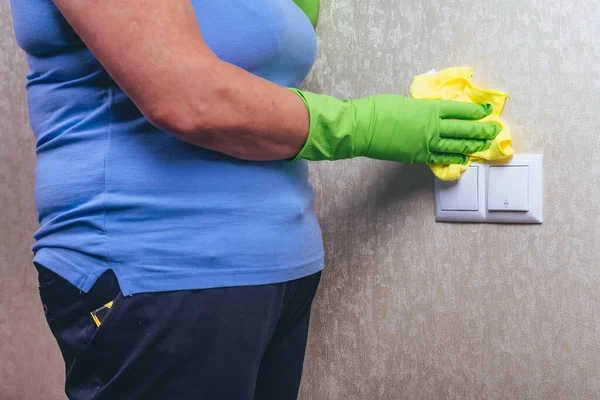 Cleaning in the house. A girl in green gloves wipes the electricity switch with a yellow cloth. cleaning from dust. Cleaning of a surface. Blue T-shirt and experienced worker.