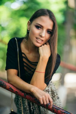 Tatlı esmer gülümsüyor ve kameraya bakıyor, ellerini tırabzana koyuyor, boruya. Güzel kahverengi gözler, siyah ceket. Yeşil doğa ve sakar kadın..