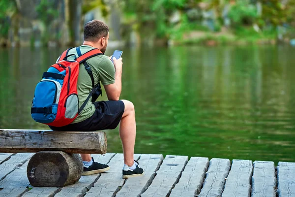 Tourist Backpack His Back Sits Tree Bench Lake Looks Phone — Stock Photo, Image