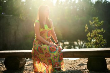 Kız doğada güzel bir gün batımı ışığıyla bir bankta oturuyor. Romantik atmosfer. Parlak ve uzun yaz elbisesi. sihirli ışıklandırma.
