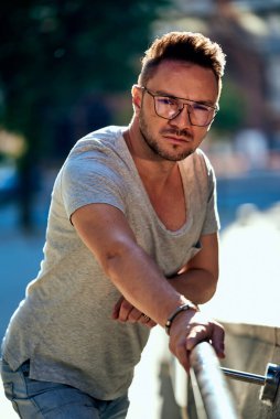 Gözlüklü şık bir adam kameraya bakar, tırabzana tutunur ve düşünür. güçlü erkek bakışları. Açık renk kot ve şık gri bir tişört. Güzel güneş ışığı..