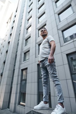 Gözlüklü şık bir adam gri bir ofis binasının yakınına bakar, elleri ceplerinde, tam yükseklikte. güçlü sert erkek bakışları. şık bir gri tişört ve açık renk kot pantolon. Şehirde portre.