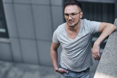 Gözlüklü şık bir adam ofis binasının yakınındaki kameraya bakıp kendini beton bir çite sarmış. güçlü sert erkek bakışları. şık bir gri tişört ve açık renk kot pantolon. Şehirde portre.