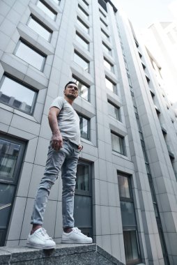 Gözlüklü şık bir adam gri bir ofis binasının yakınına bakar, elleri ceplerinde, tam yükseklikte. güçlü sert erkek bakışları. şık bir gri tişört ve açık renk kot pantolon. portre.