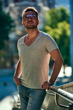 Gözlüklü şık bir adam yukarı bakar, tırabzana tutunur ve düşünür. güçlü erkek bakışları. Açık renk kot ve şık gri bir tişört. Güzel kontollü güneş ışığı. yaz.