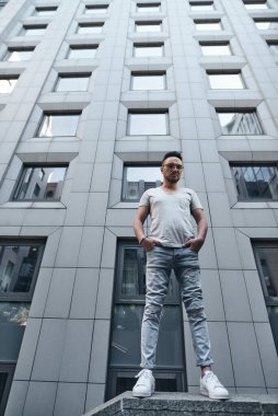 Gözlüklü şık bir adam, aşağı bakıyor, ofis binasının yakınında, elleri ceplerinde, tam yükseklikte. güçlü sert erkek bakışları. şık bir gri tişört ve açık renk kot pantolon. Şehirde portre.