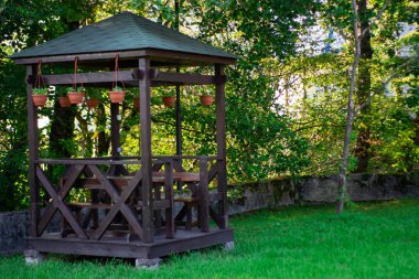 Outdoor wooden gazebo over summer landscape background. Wood brown arbour on green lawn. Summertime lush foliage pine trees blue cloudy sky sunny day nobody. clipart